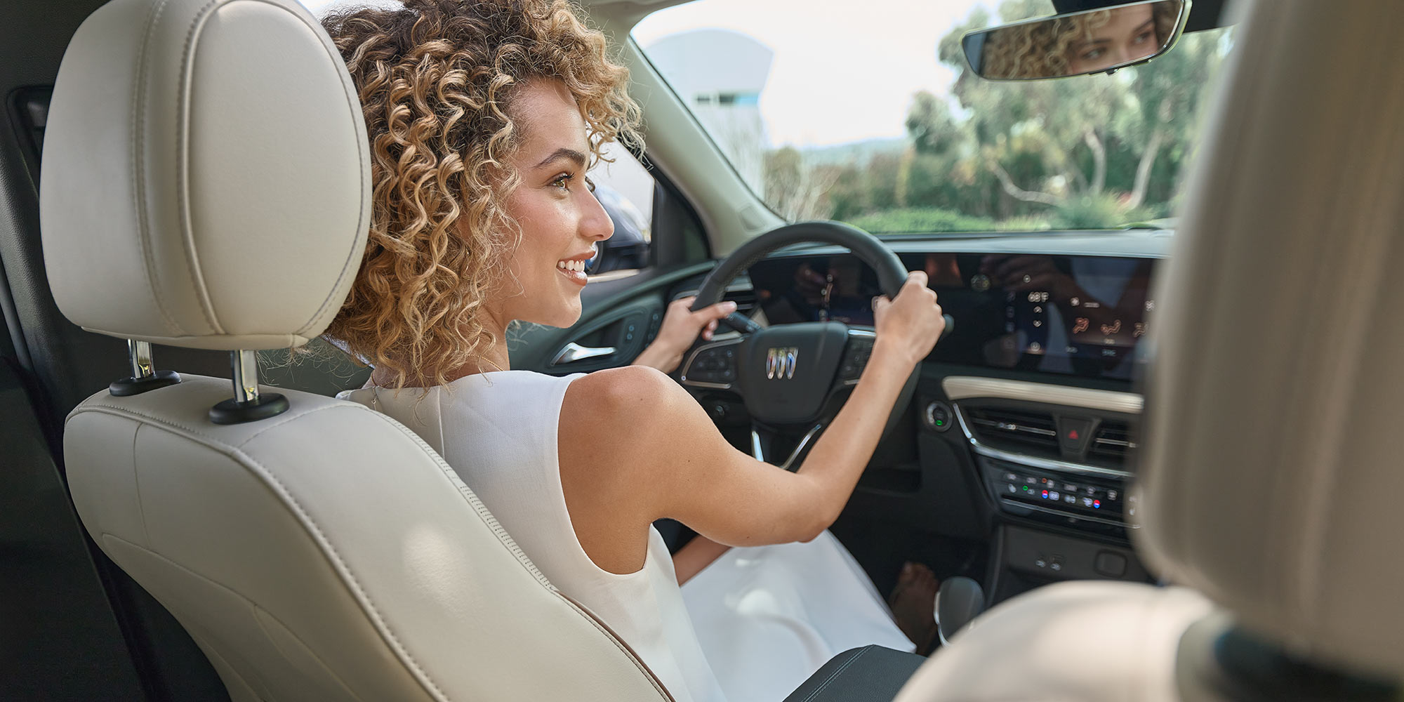 A Smiling Woman with Curly Blond Hair Driving a Buick Envision Interior with Light Colored Seats and a Digital Dashboard