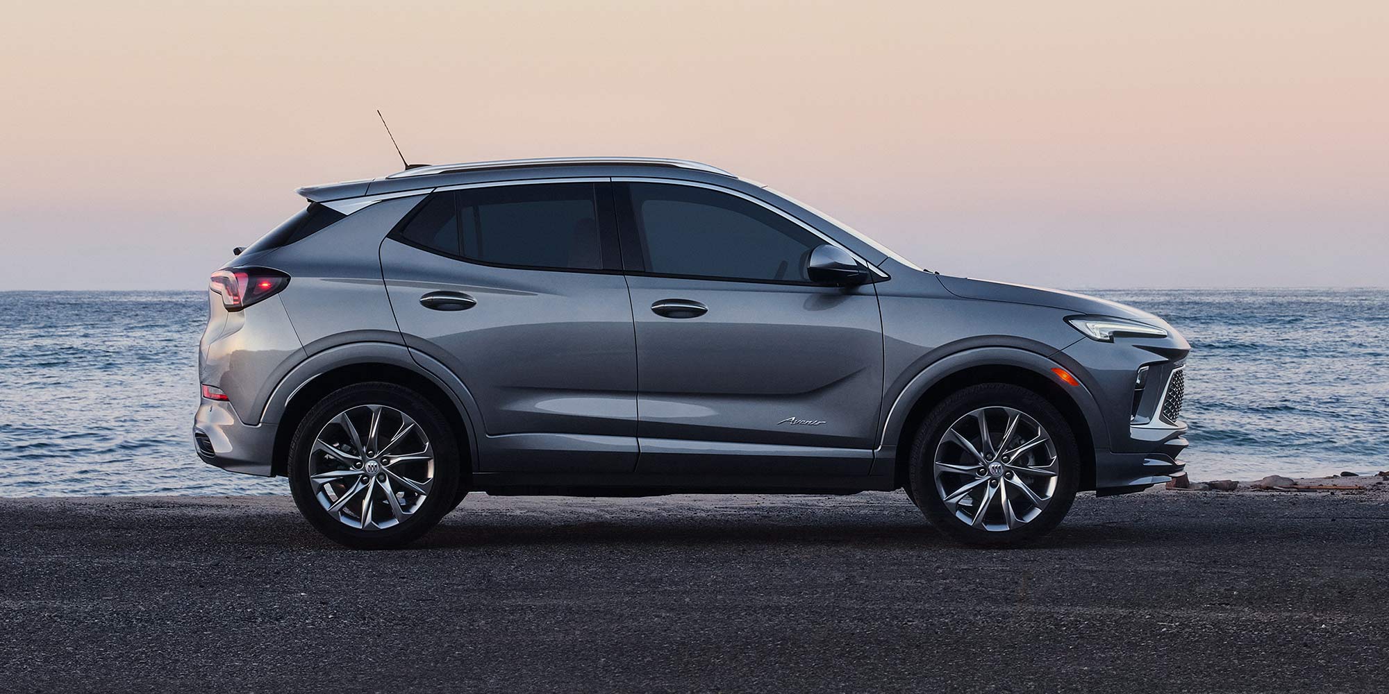 A Side Profile View of a Gray Buick Encore GX SUV Parked on the Asphalt Road by the Ocean at Sunset
