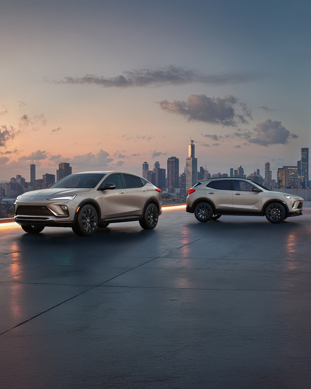 The 2026 Envista Sport Touring SUV and the 2026 Encore GX Sports Touring SUV both in the Color Ceramic Parked on a Parking Garage Rooftop Overlooking a City Skyline