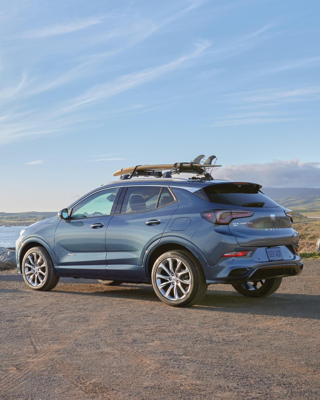 Rear Three-Quarters View of a Buick Encore GX Avenir Luxury SUV Parked Overlooking a Large Body of Water with a Surfboard Attached to the Top of the Vehicle