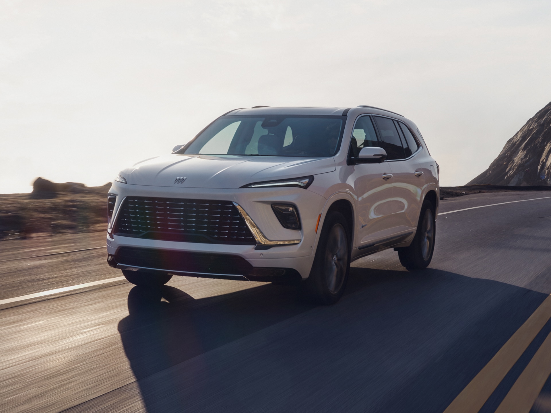 A White Buick Envision Avenir SUV Driving Up a Winding Road with Rocky Mountains Under a Bright Sky