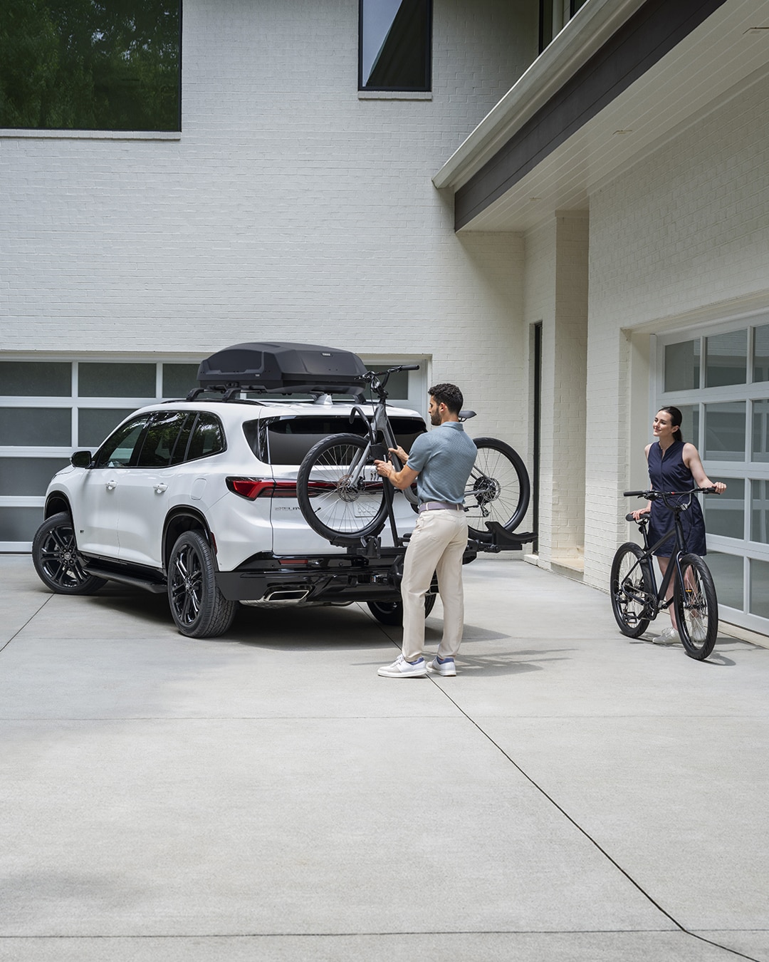 A White Buick SUV in a Driveway with a Man Loading a Bicycle onto the Rear Rack and a Woman Stands Nearby with Another Bicycle