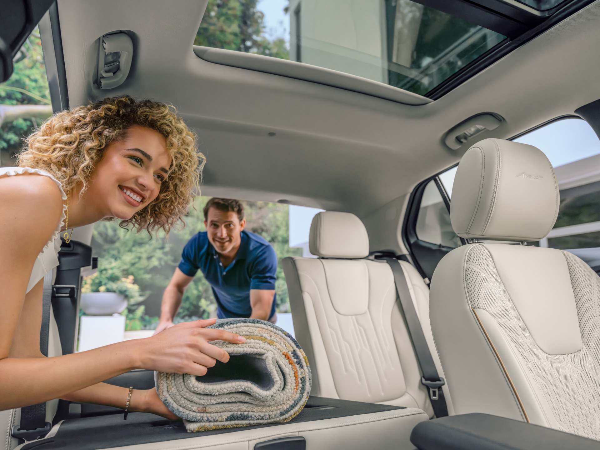 A Smiling Woman Loads a Rolled Rug into the Open Trunk of a White Buick SUV, Which Features Light-Colored Leather Seats and a Sunroof While a Man Watches in the Background