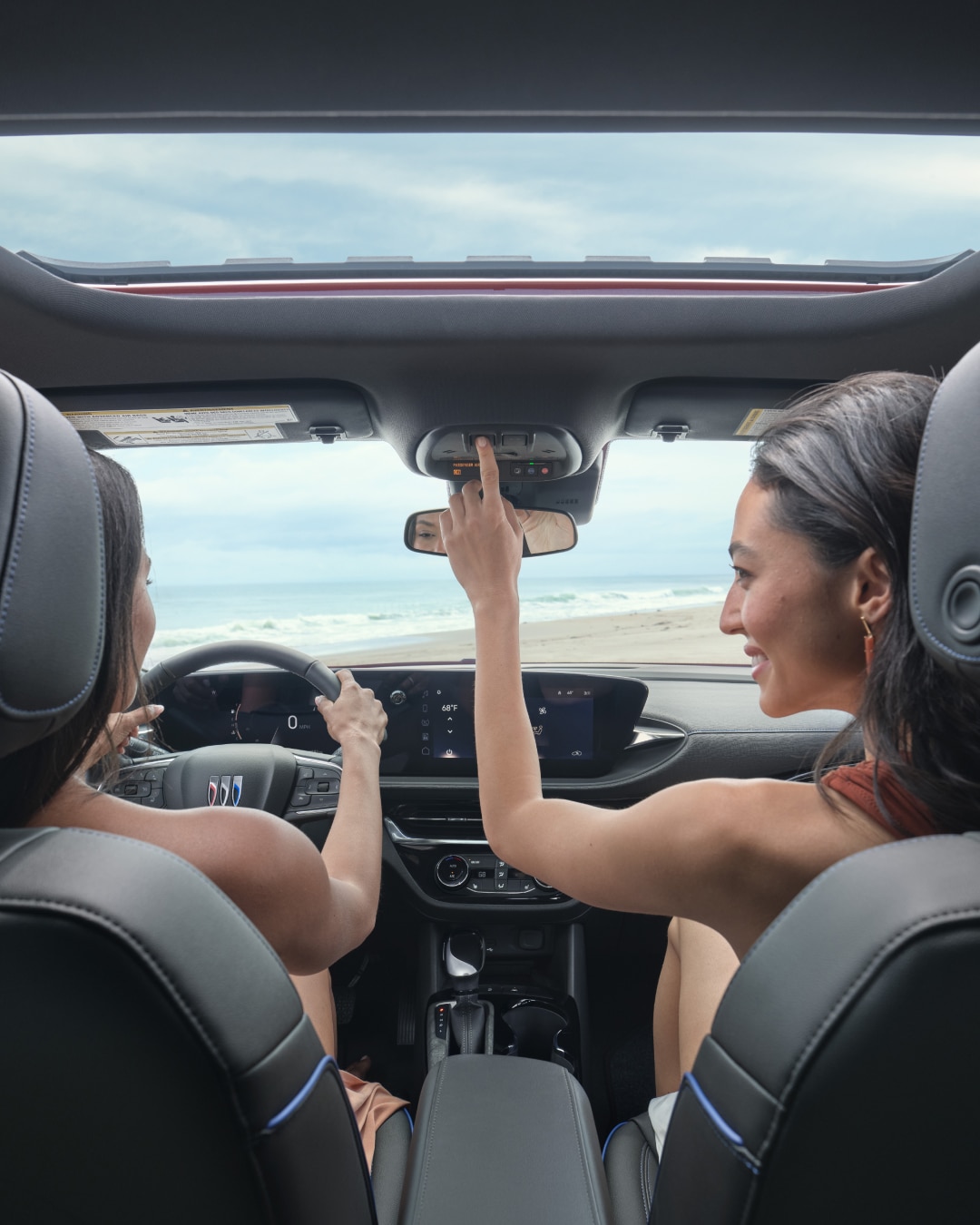 Two Women Drive a Buick Along the Ocean One Steers with the Buick Logo and the Other Adjusts the Overhead Console