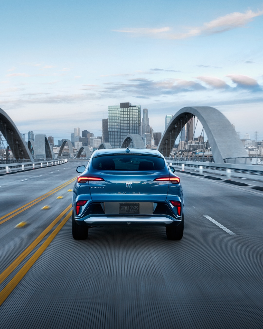 A Rear View of a Blue Buick Envista SUV Driving on a Bridge Toward a City Skyline at Dusk