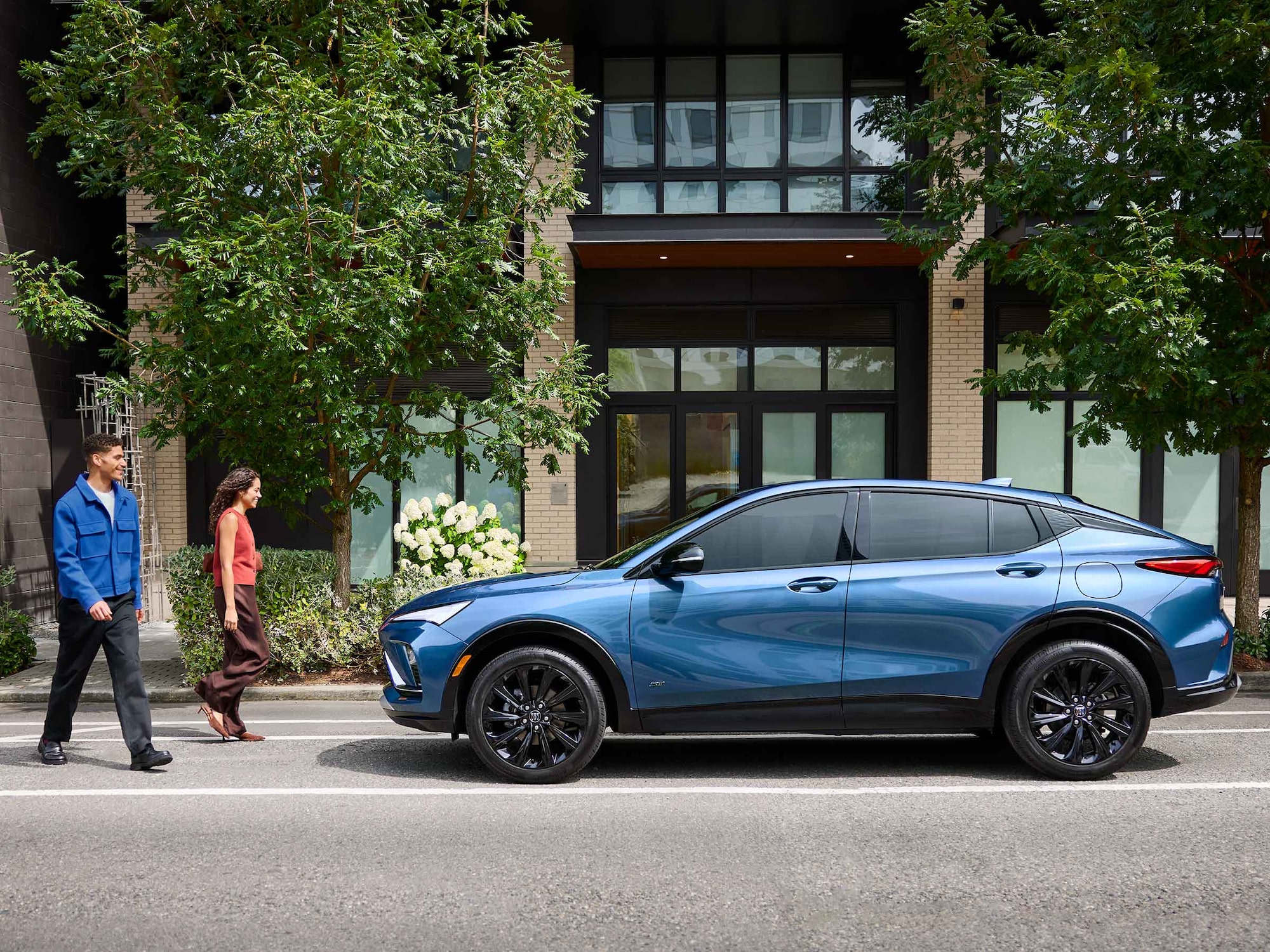 A Metallic Blue Buick Envista Parked on a City Street with a Couple Walking in Front of it and Green Foliage in the Background
