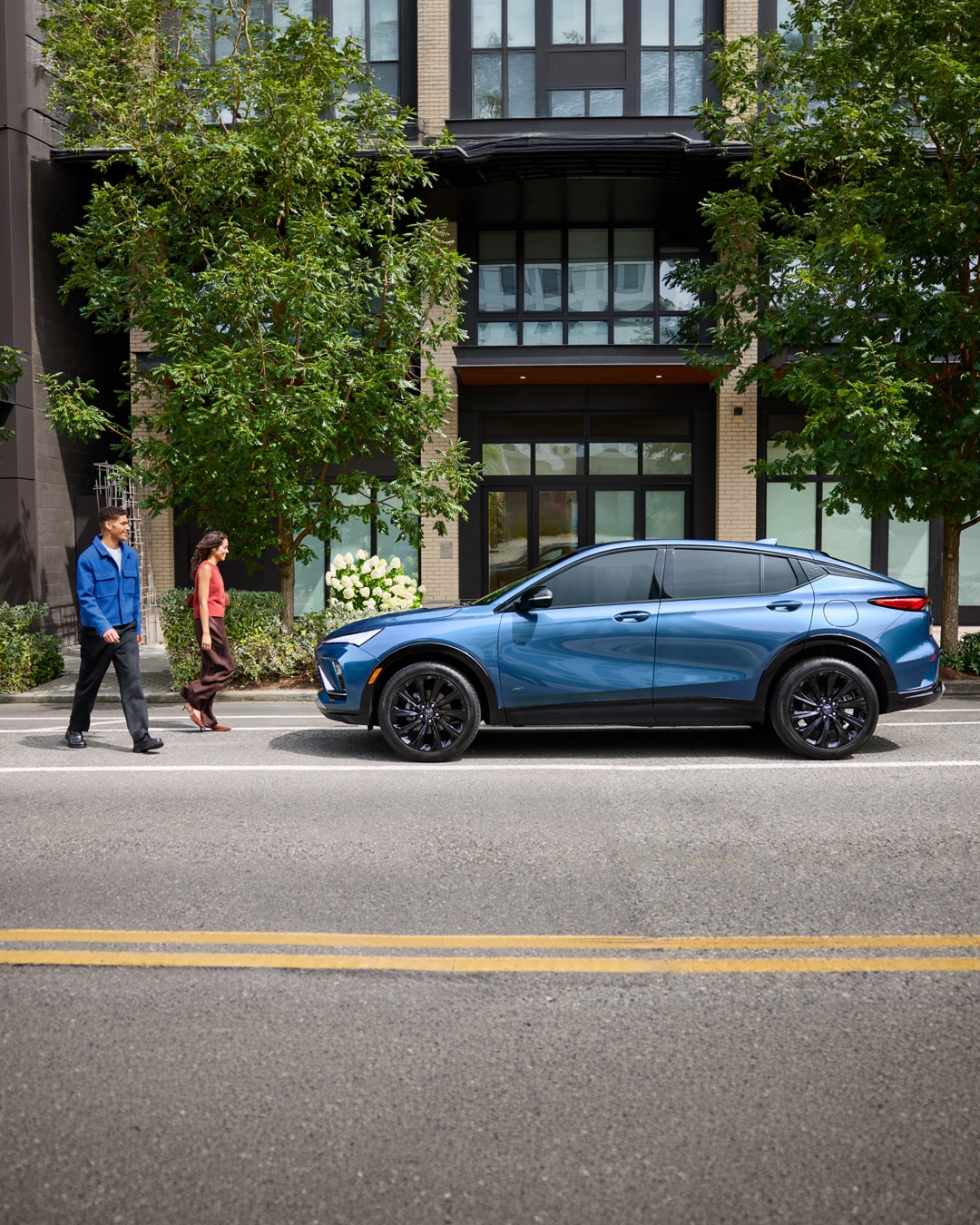 A Metallic Blue Buick Envista Parked on a City Street with  Green Foliage in the Background and a Couple Walking in Front of it