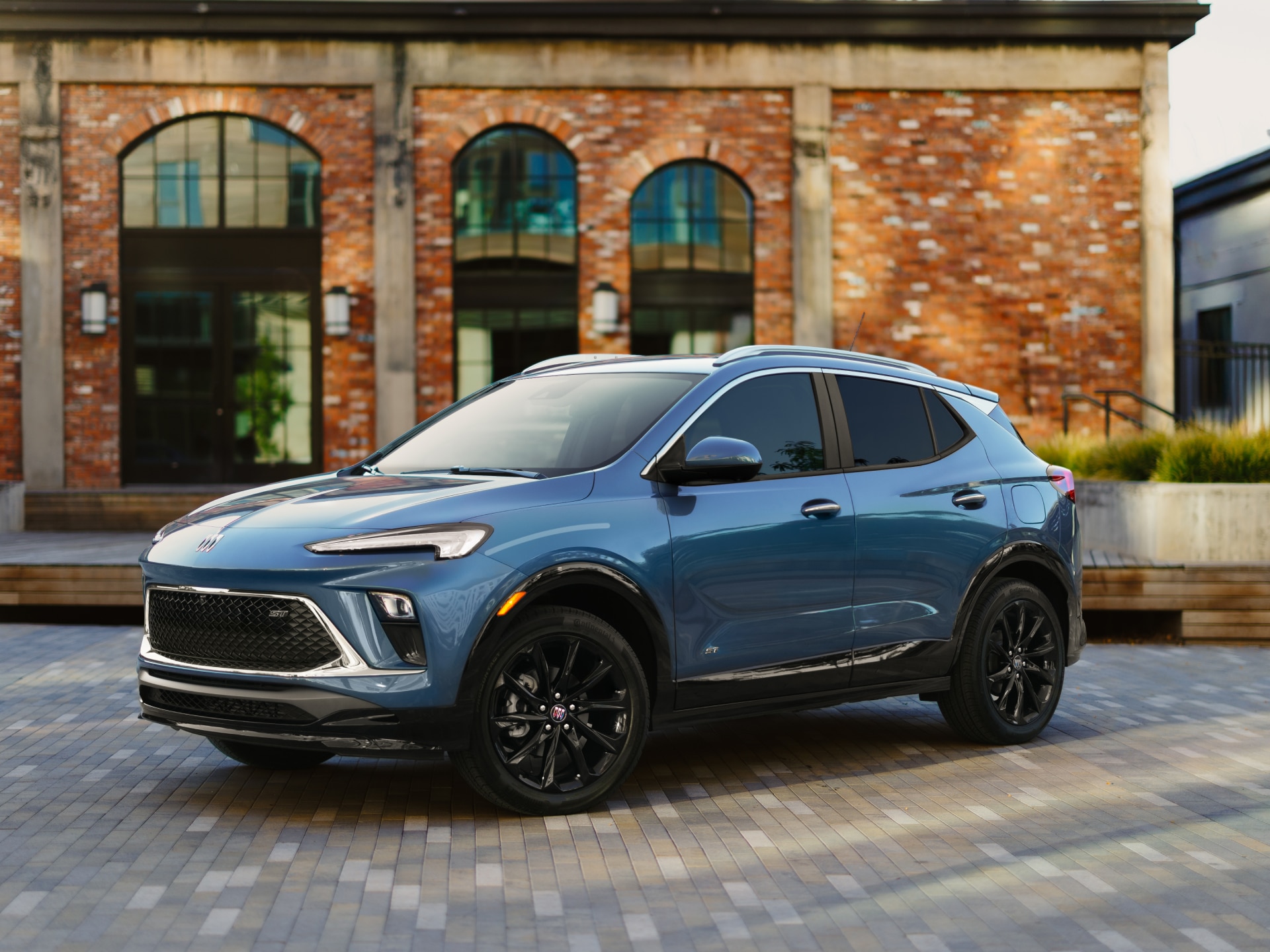 A Metallic Blue Buick Encore GX Parked on a Patterned Brick Walkway in Front of an Old Brick Building with Large Windows