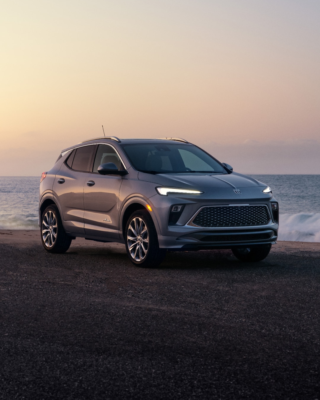 A Three-Quarters View of a Silver Buick Encore GX Avenir Parked on a Beach at Sunset
