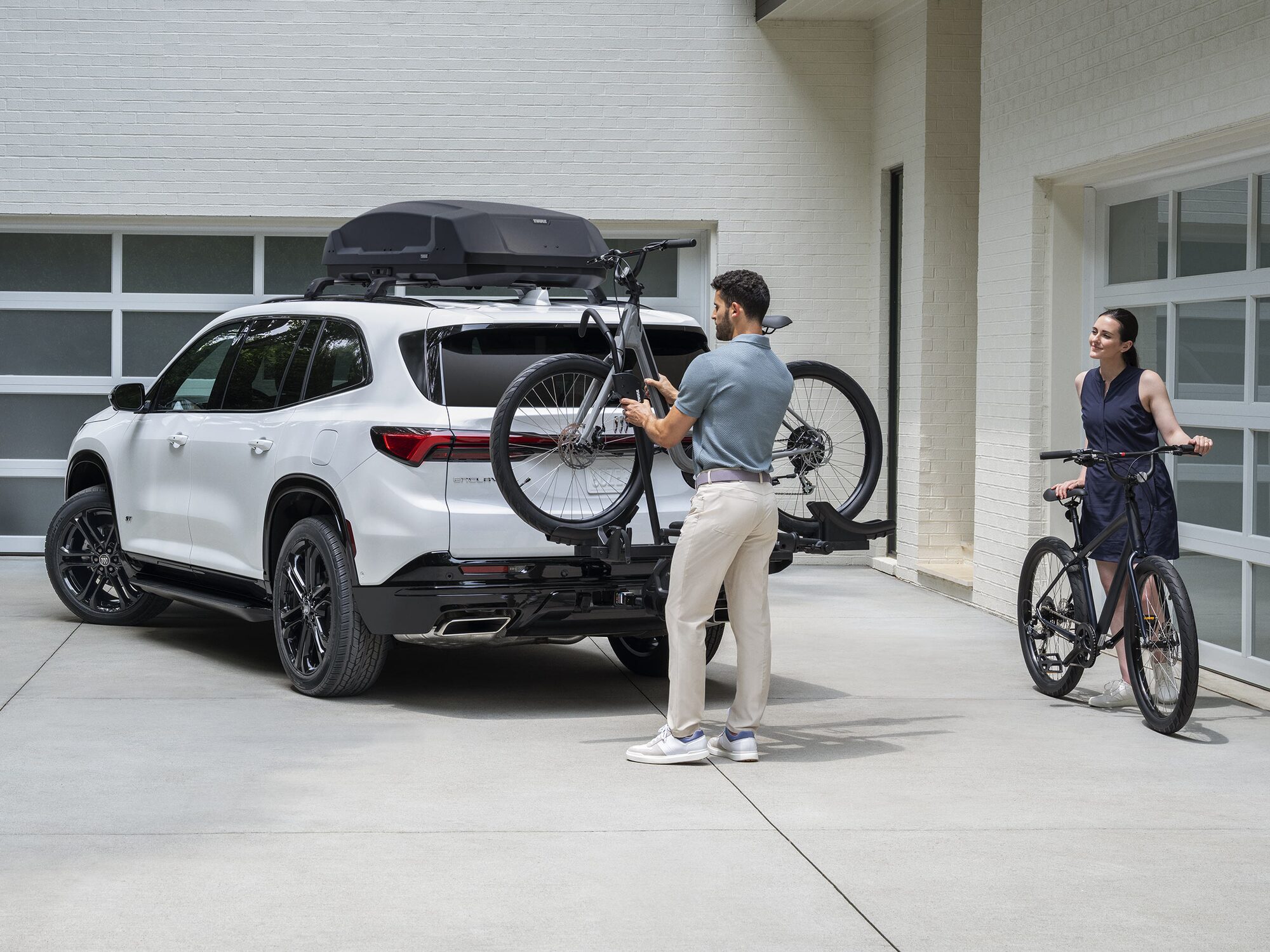 A White Buick SUV with a Roof Rack and Bike Carrier Is Parked in a Driveway a Man Loads a Bicycle onto the Rear Rack While a Woman Stands Nearby with Another Bicycle