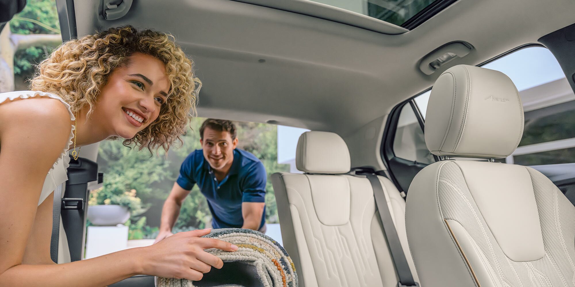 A Smiling Woman Loads a Rolled Rug into the Open Trunk of a White Buick SUV, Which Features Light-Colored Leather Seats and a Sunroof While a Man Watches in the Background 