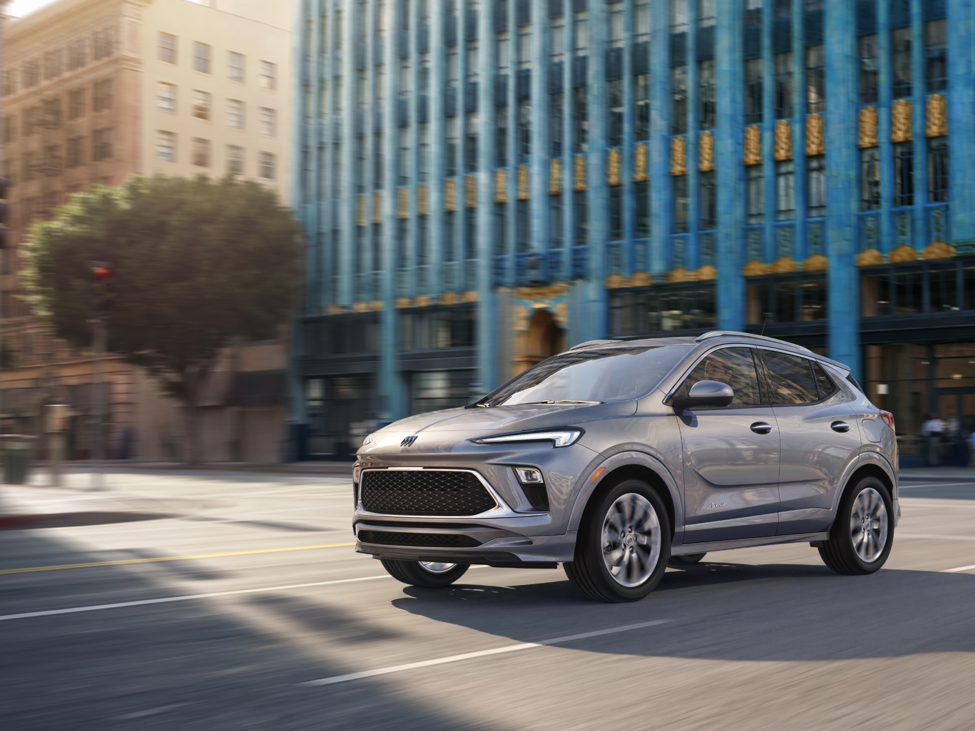 A Gray Buick Envista SUV Drives on a City Street in Front of Modern Buildings