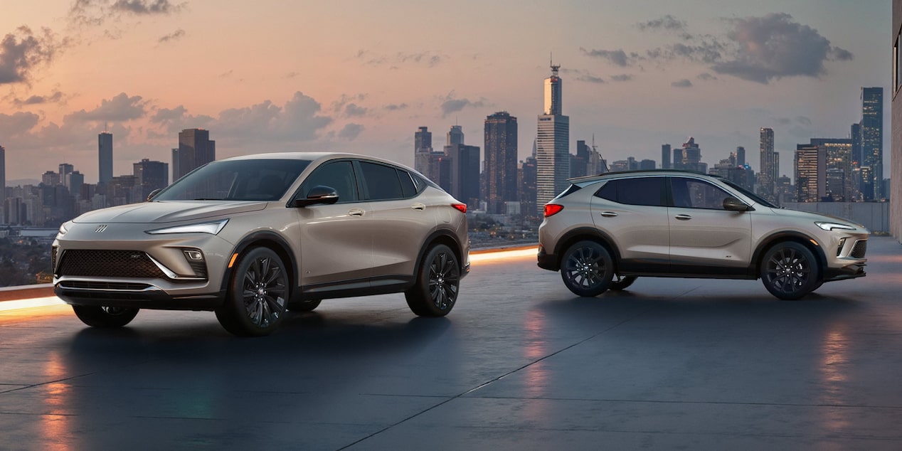 The 2026 Envista Sport Touring SUV and the 2026 Encore GX Sports Touring SUV both in the Color Ceramic Parked on a Parking Garage Rooftop Overlooking a City Skyline