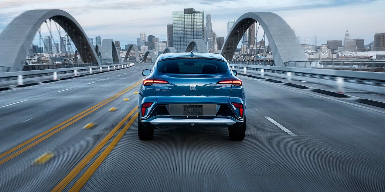 A Rear View of a Blue Buick Envista SUV Driving on a Bridge Toward a City Skyline at Dusk