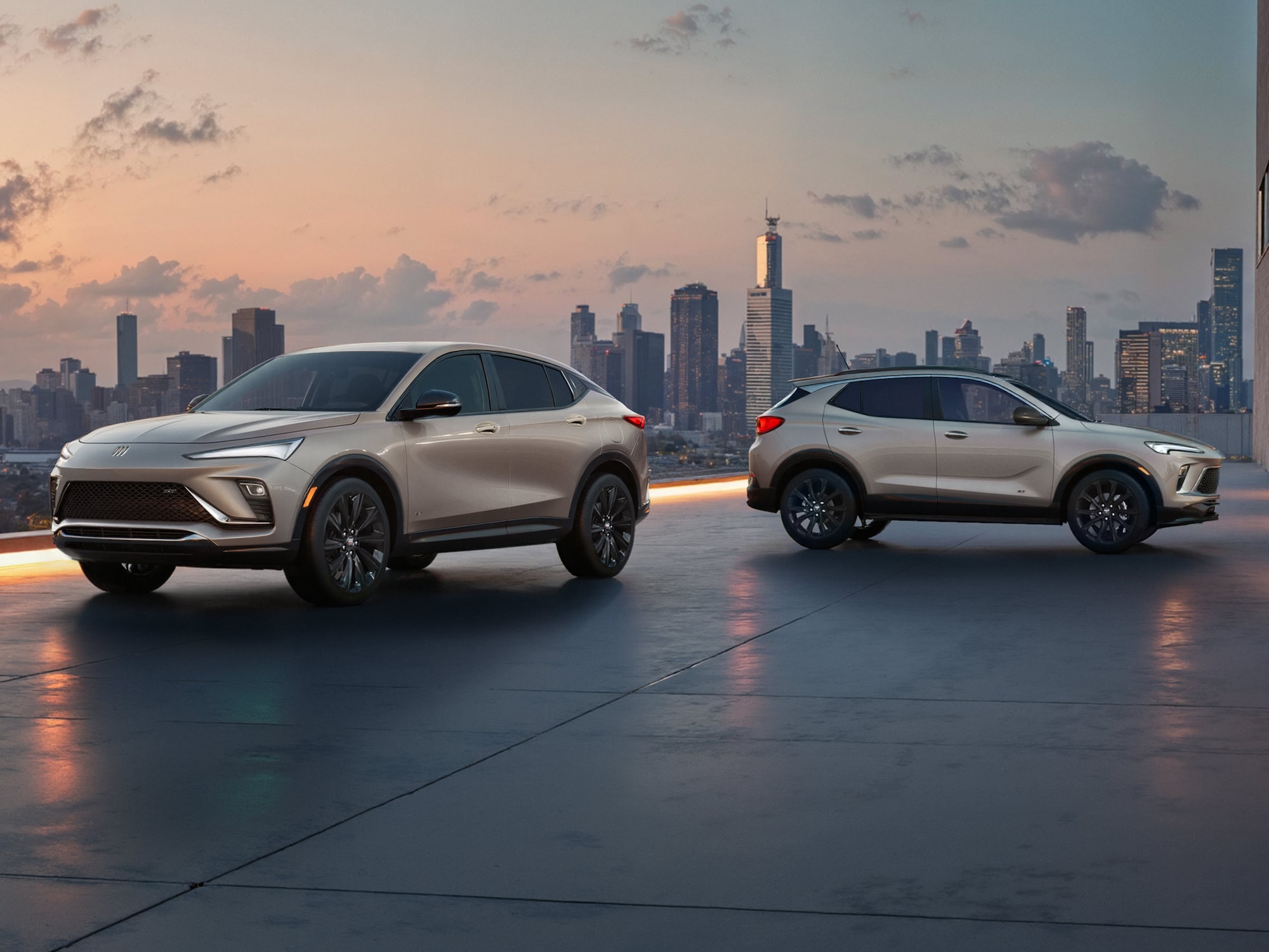 The 2026 Envista Sport Touring SUV and the 2026 Encore GX Sports Touring SUV both in the Color Ceramic Parked on a Parking Garage Rooftop Overlooking a City Skyline 