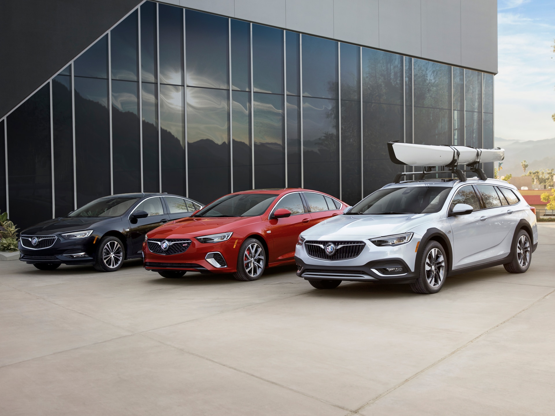A Black Buick Sedan, a Red Buick Regal GS Sedan, and a Light Gray Buick Regal TourX Wagon with a Kayak on its Roof Rack are Parked in Front of a Modern Building