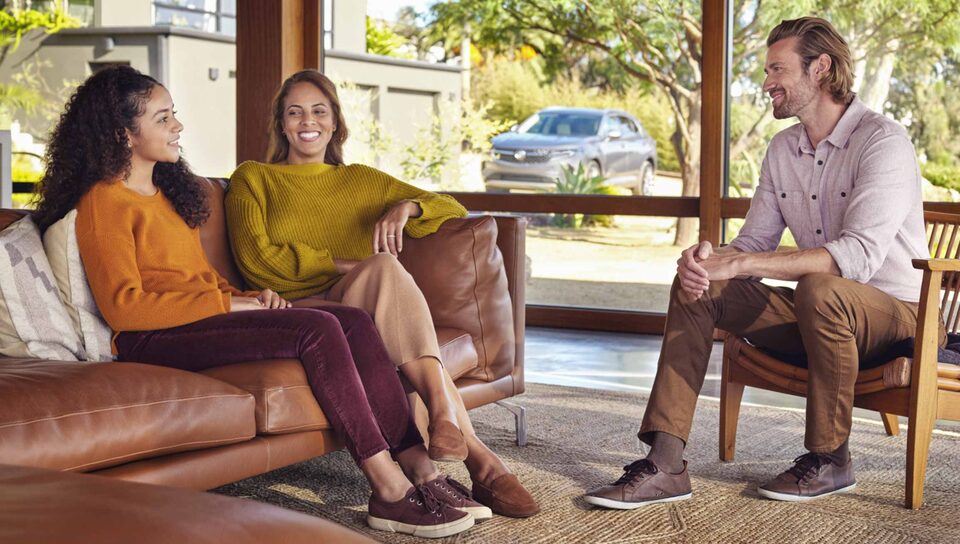 A woman, man and young woman sit inside the living room of a modern home. The all—new Buick Avenir sits in the driveway and is visible from the window