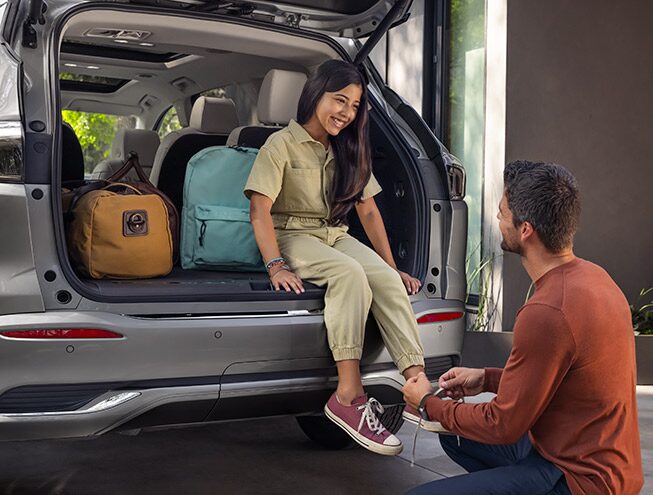 A Man Ties His Daughter's Shoe As She Sit in the Trunk of Their Buick Encore