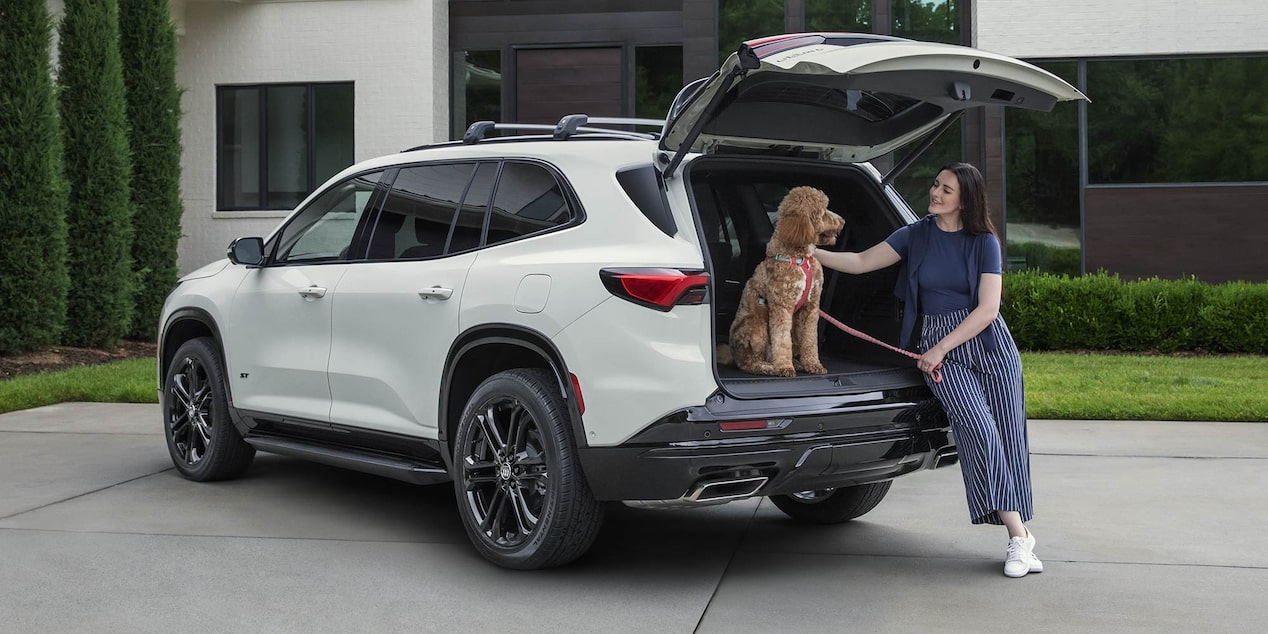 A Woman Sitting Beside the Open Liftgate of a White Buick Enclave SUV Petting a Brown Poodle Wearing a Red Harness