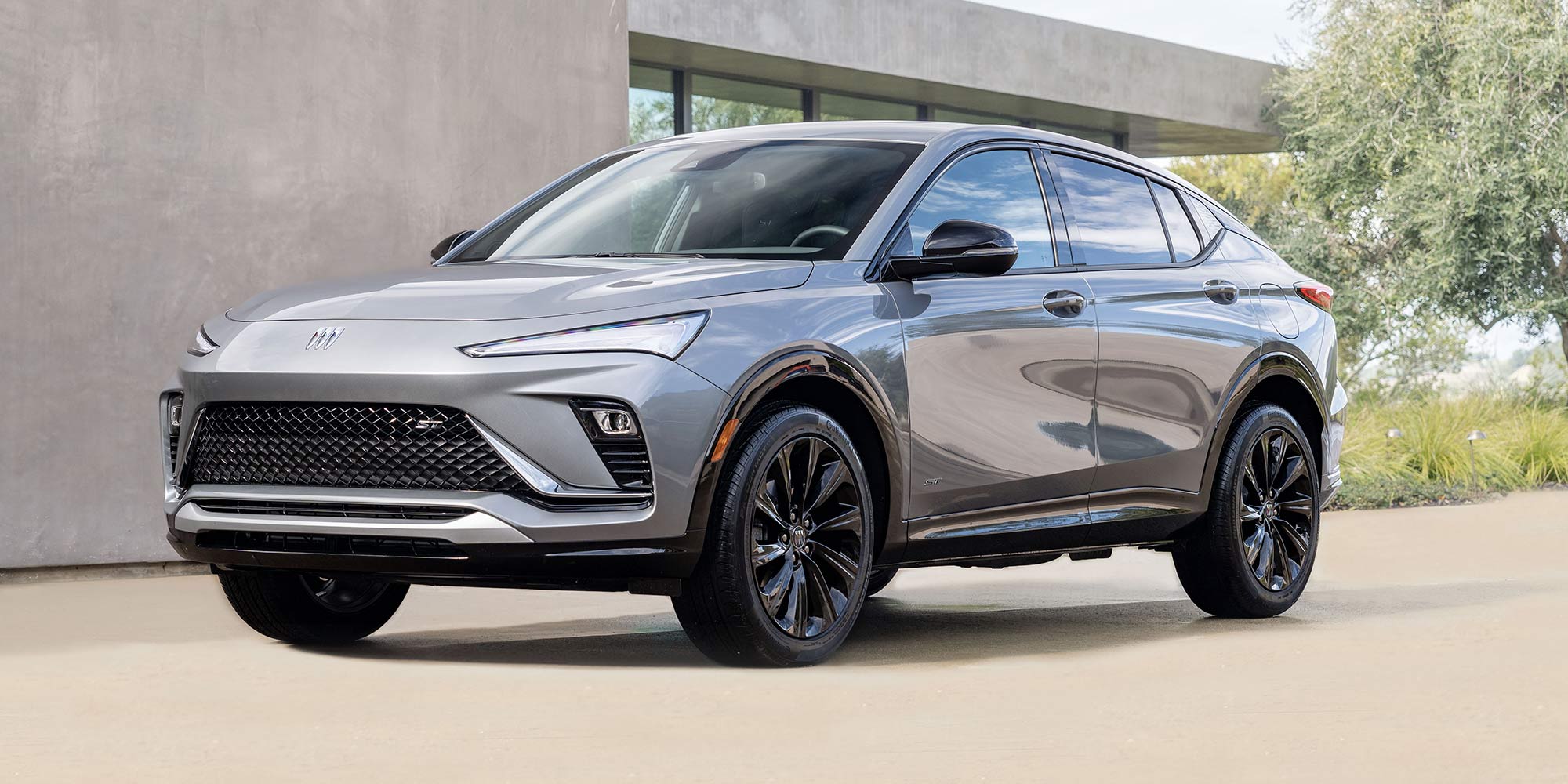 A Front Quarter View of a Sleek Gray Buick Envision Avenir SUV with Black Wheels Parked in Front of a Modern Concrete Building