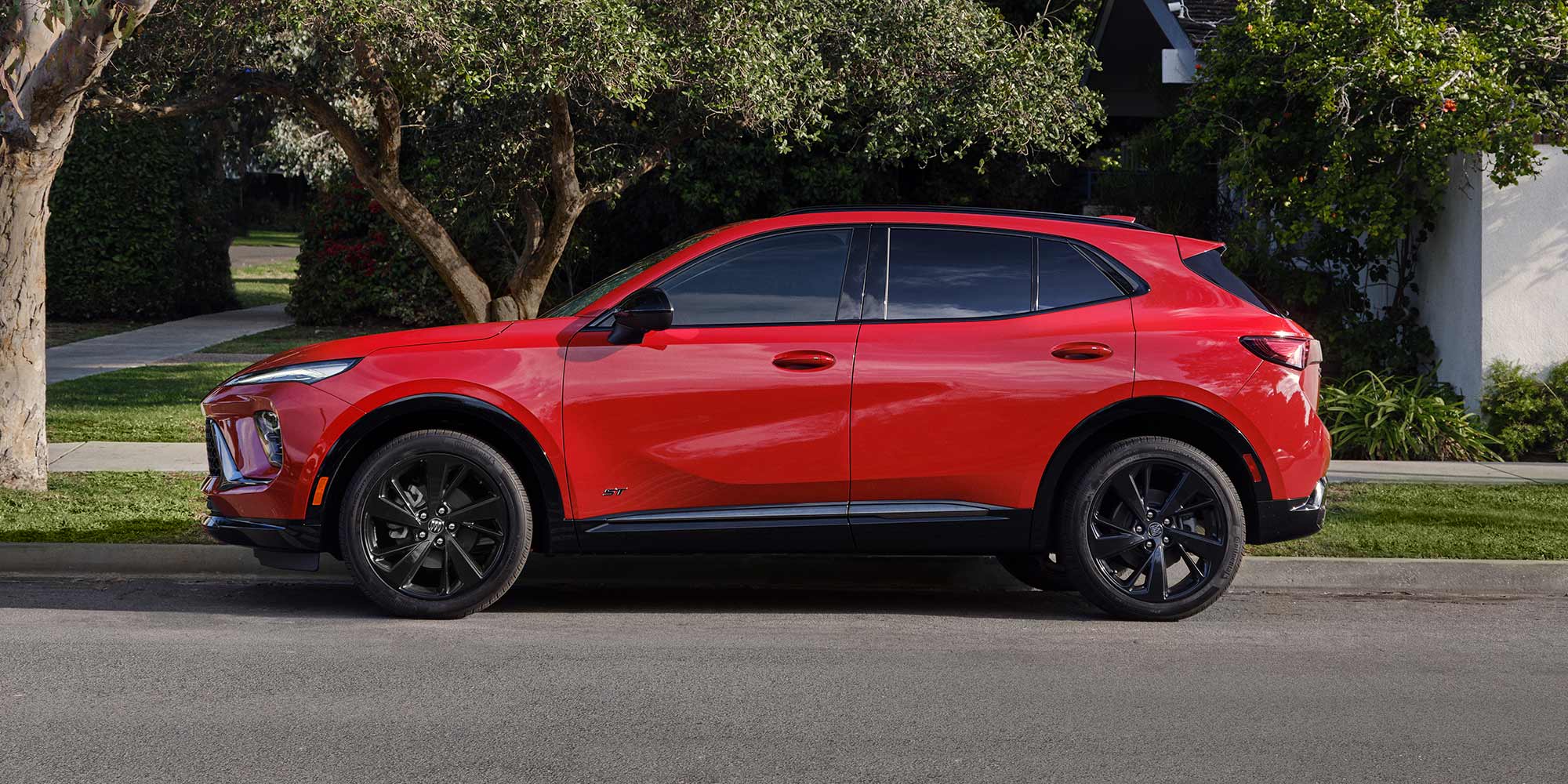 A Side Profile of a Vibrant Red Buick Envision SUV with Black Wheels Parked on a Street Next to a Sidewalk and Green Trees