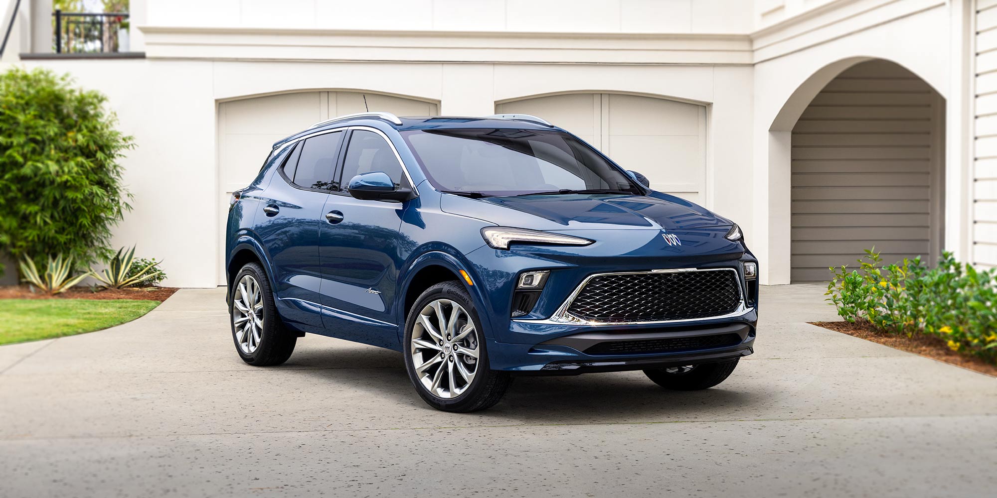 A Front Quarter View of a Shiny Dark Blue Buick Encore GX SUV with Silver Wheels Parked on a Paved Driveway in Front of a White House with Garage Doors