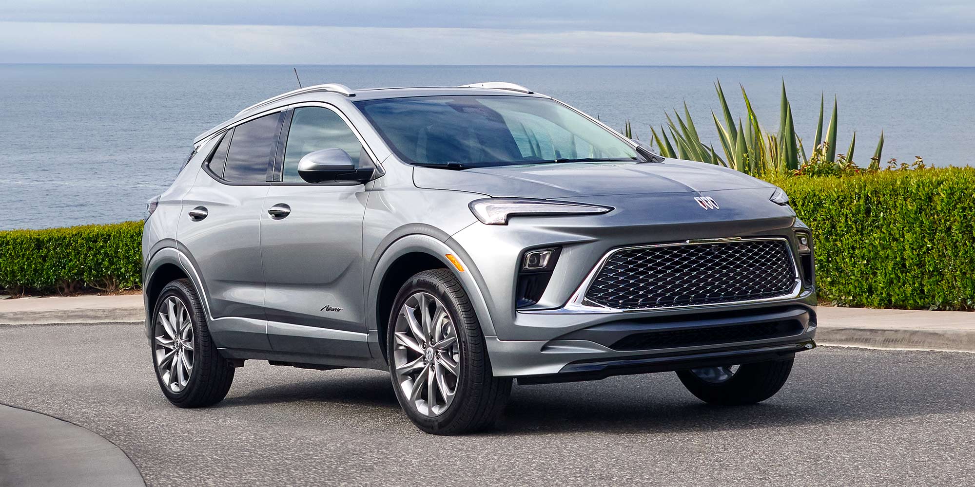 A Silver Buick Envista Parked on an Asphalt Road with the Ocean and Green Plants in the Background 