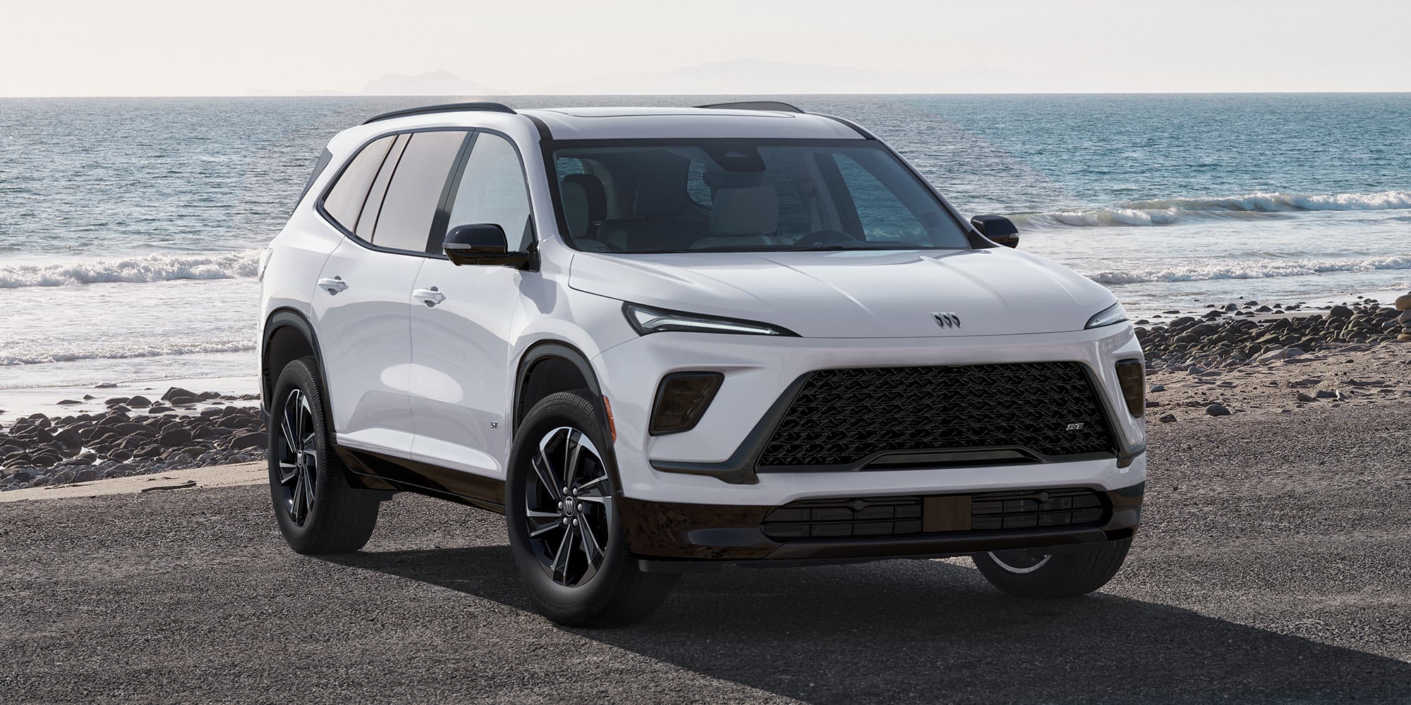 A White Buick Envista Parked on a Rocky Beach with the Ocean and a Hazy Sky in the Background 