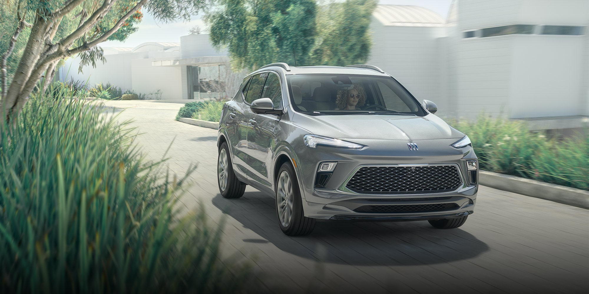 A Gray 2026 Buick Encore GX SUV Parked in a Driveway of a Modern House with Green Foliage