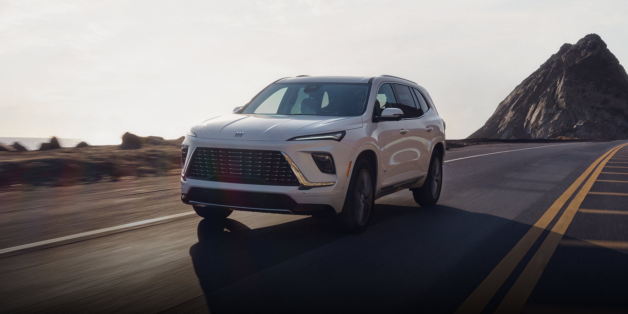 A White 2026 Buick Enclave SUV Driving on a Winding Road with a Mountain and Sunset Sky in the Background