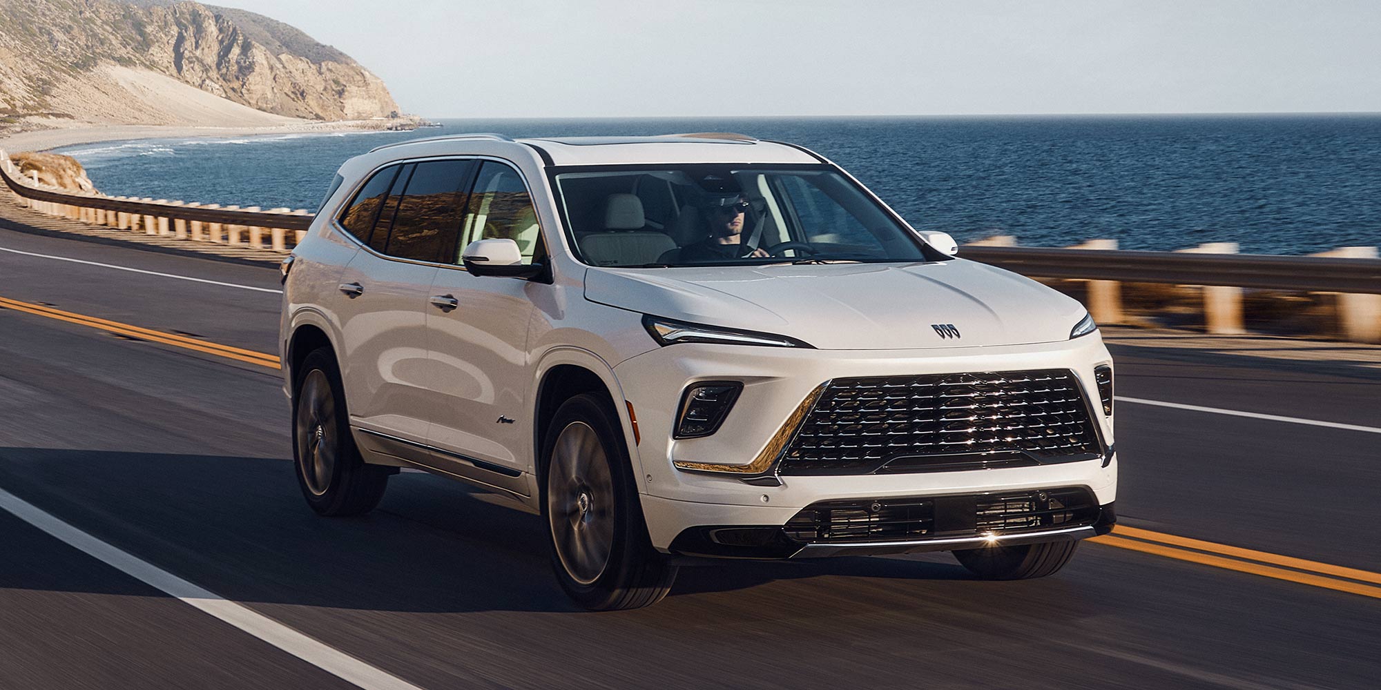 A White Buick SUV Driving on a Scenic Coastal Road With the Ocean and Cliffs in the Background on a Bright Day