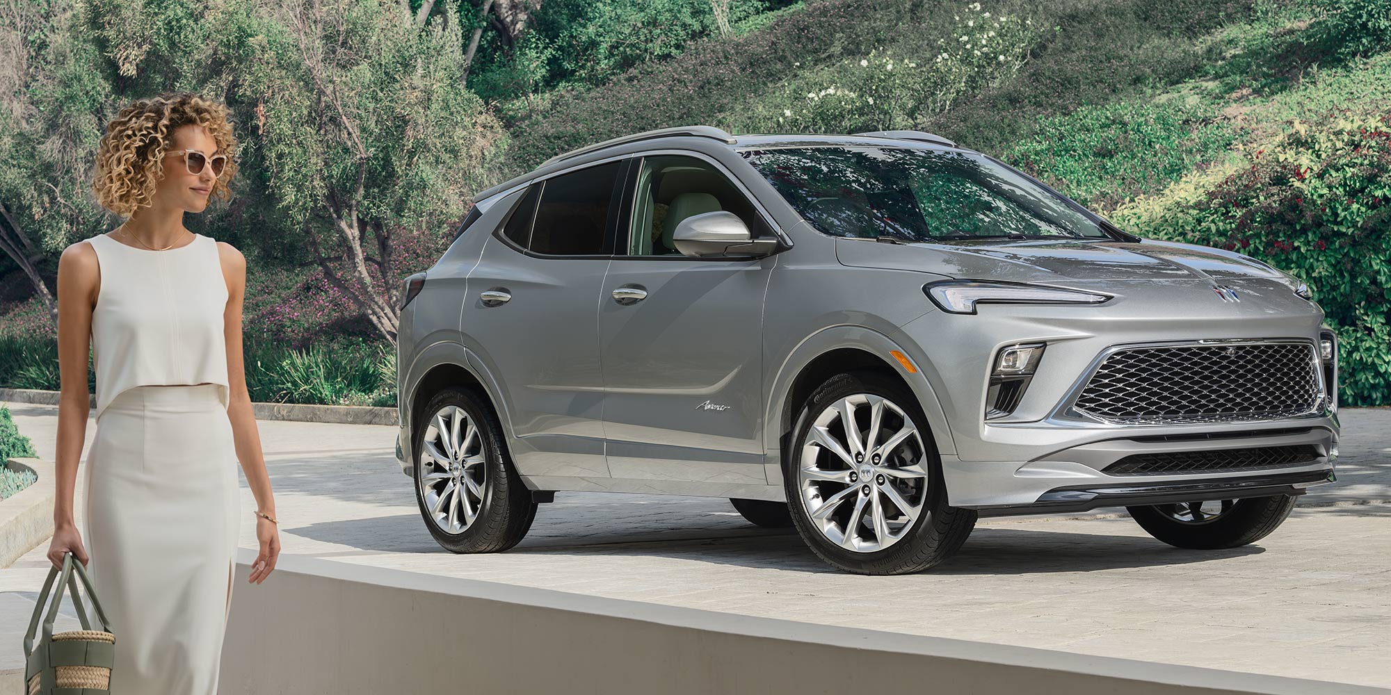 A Silver Buick Encore GX SUV Parked on a Terrace as a Woman in a White Outfit Walks Down Steps Holding a Woven Bag