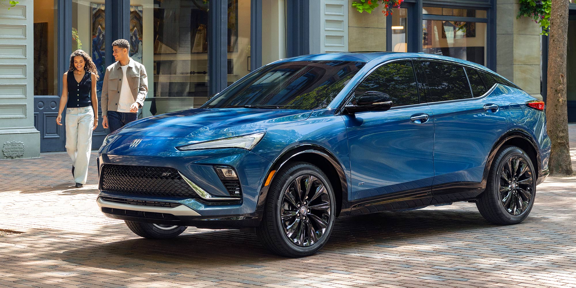 A Brilliant Blue Buick Envista ST SUV Parked on a Brick Sidewalk as Two People Walk Past a Storefront in the Background