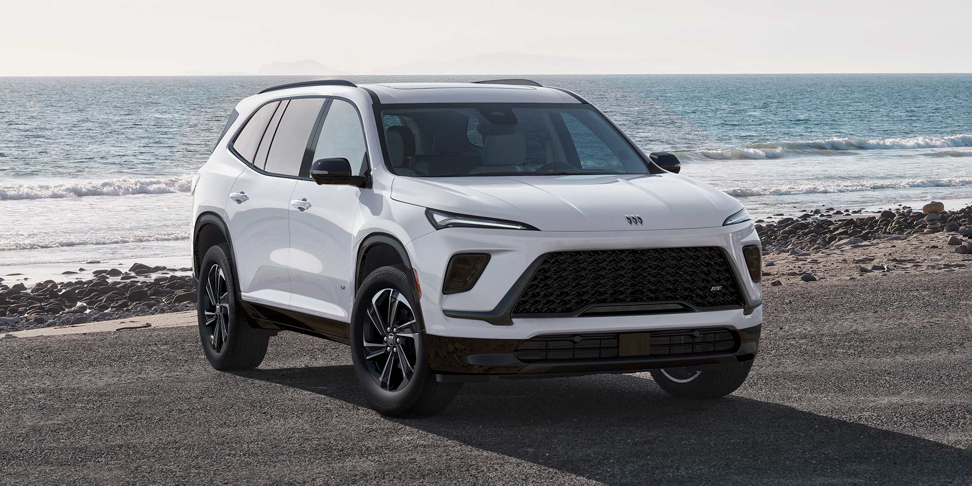 A White Buick Envision SUV Parked on a Sandy Beach with the Blue Ocean and Sky Behind It