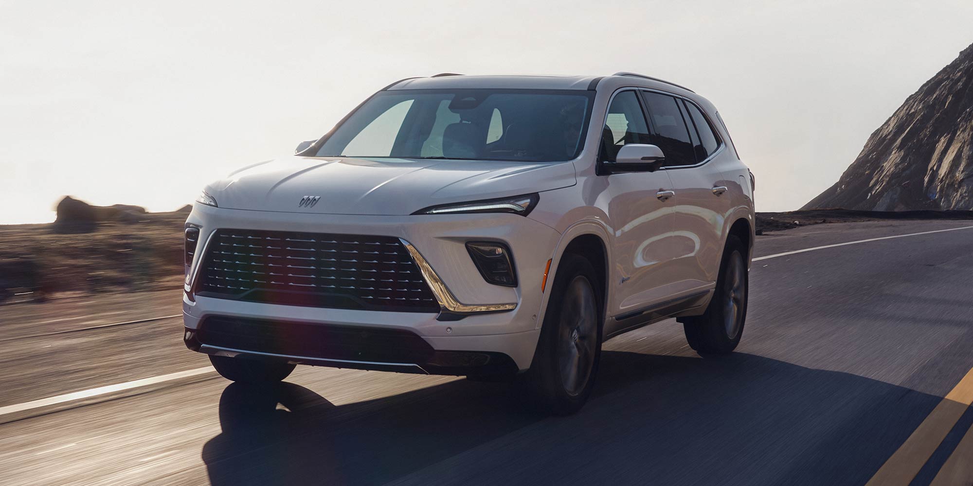 A White Buick Envision Avenir SUV Driving Up a Winding Road with Rocky Mountains Under a Bright Sky