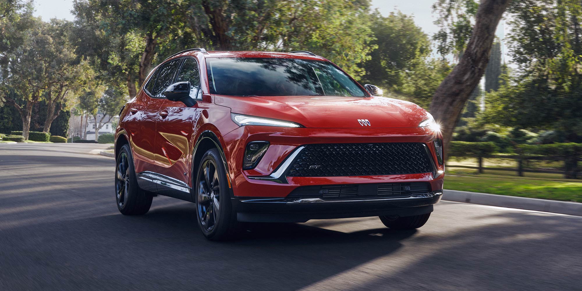 A Striking Red Buick Envista ST Driving Towards the Viewer on a Sunny Road Lined with Green Trees