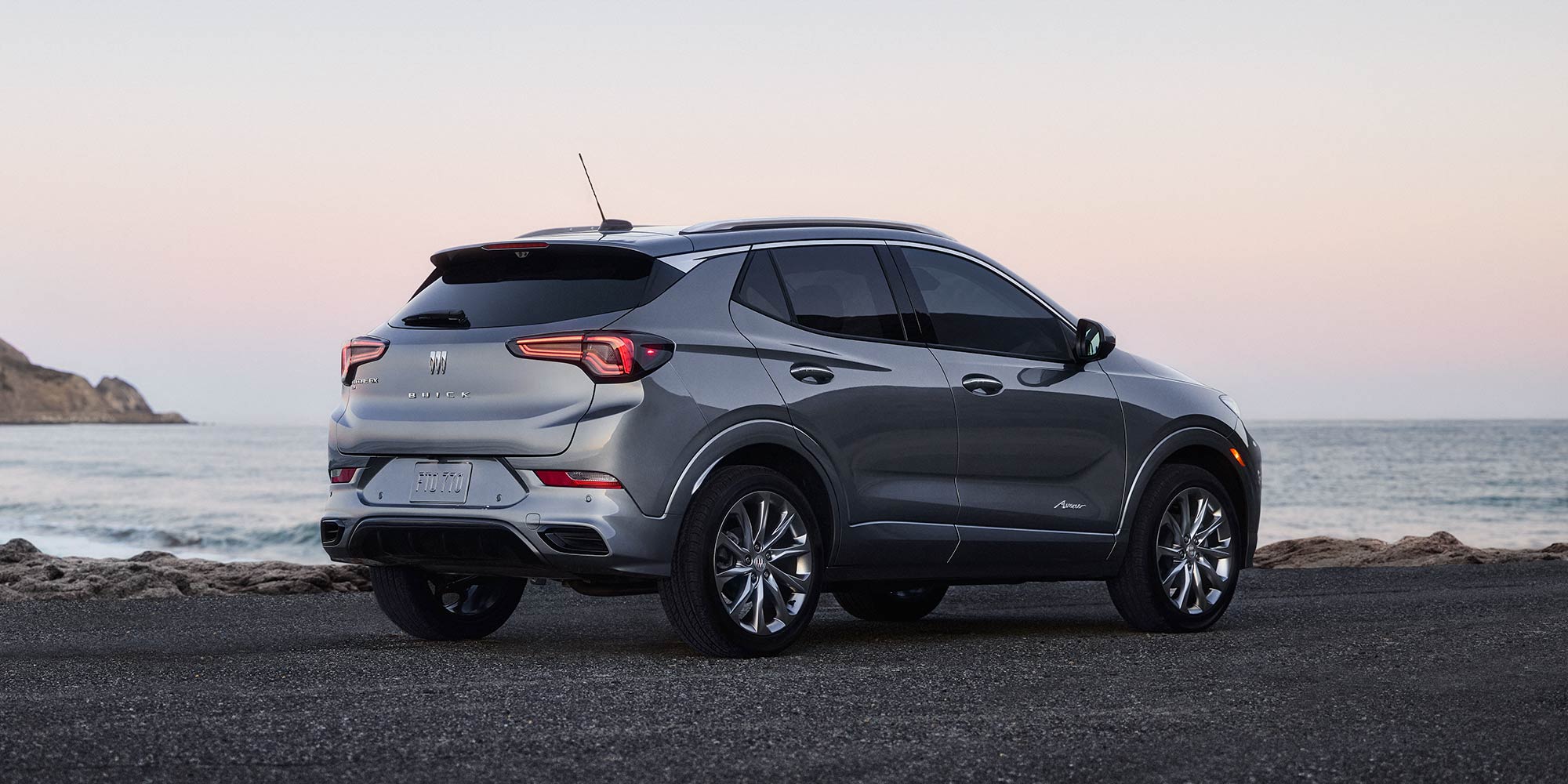 A Gray Buick Encore GX SUV From The Rear Three-Quarter View Parked By The Ocean At Sunset