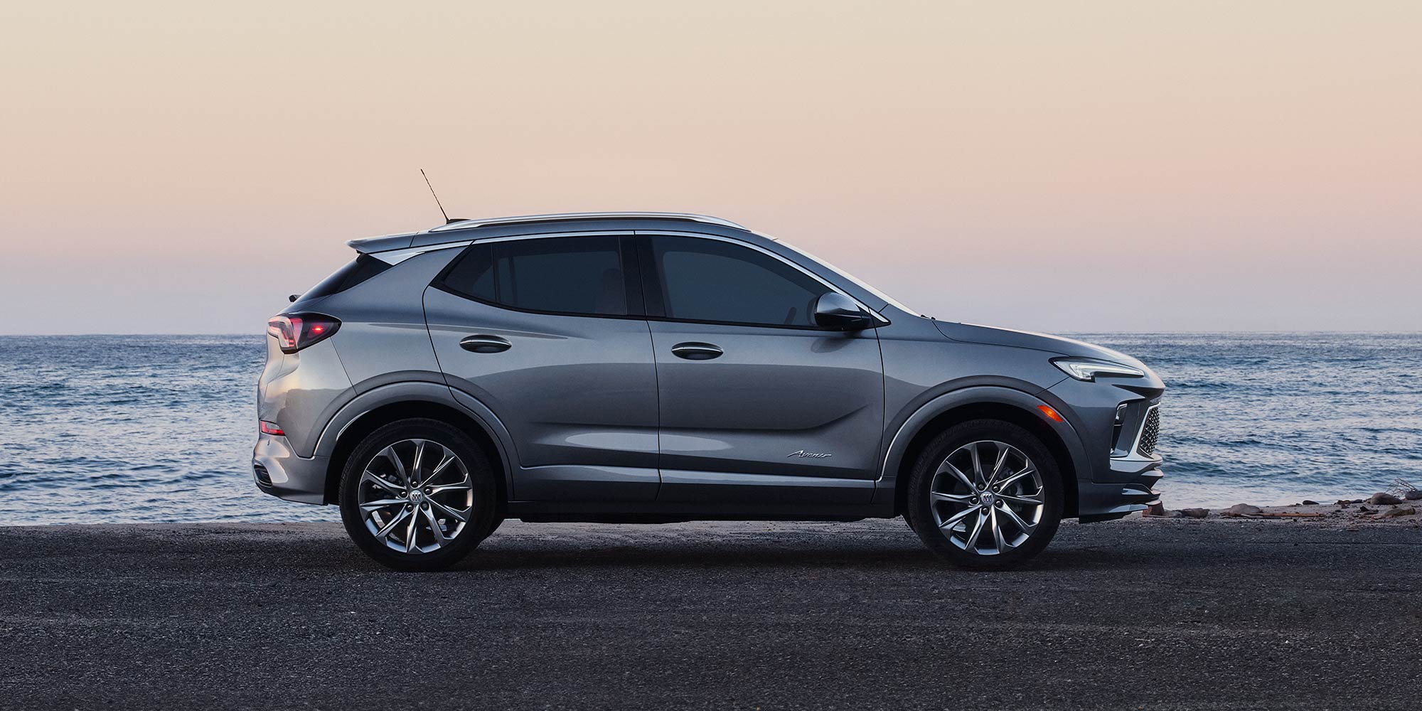 A Side Profile View of a Gray Buick Encore GX SUV Parked On the Asphalt Road By The Ocean At Sunset