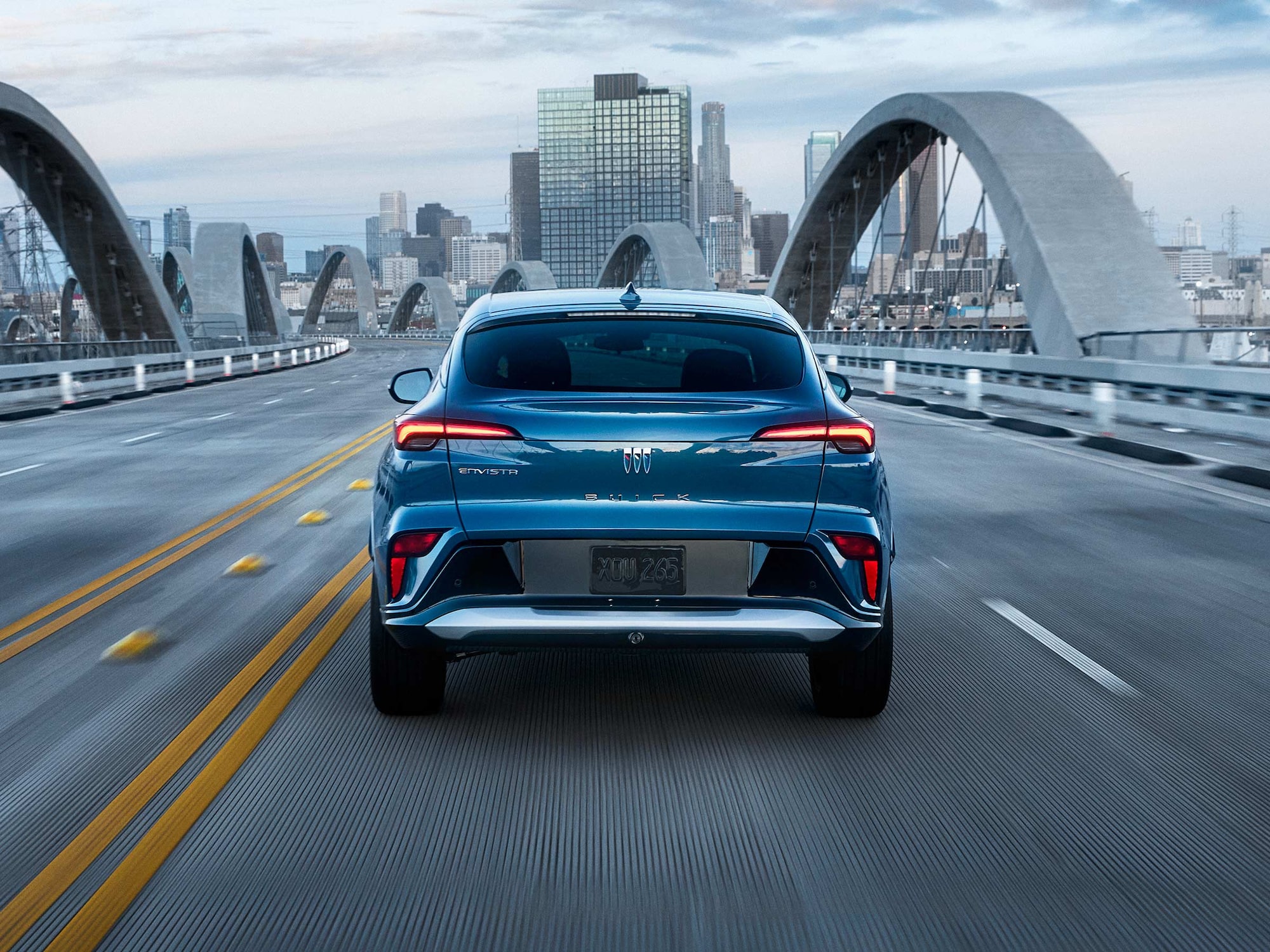 A Rear View of a Blue Buick Envista SUV Driving on a Bridge Toward a City Skyline at Dusk