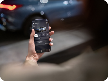 A hand holds a smartphone displaying the Buick mobile app with a car image, with a blurred Buick vehicle in the background.