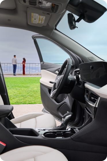 The interior view from the driver's side of a Buick, showing light-colored leather seats and an open door with a seaside view.