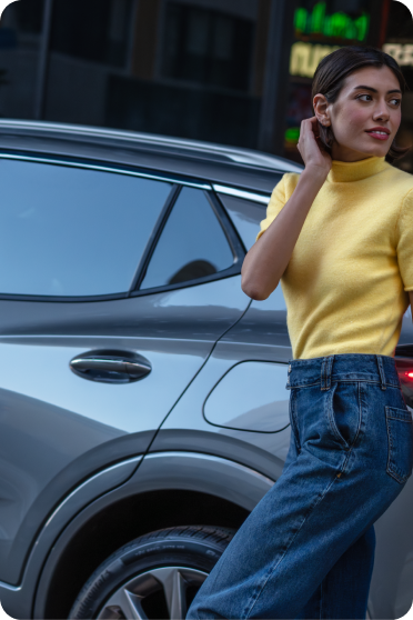 A person in a yellow top stands near a parked grey Buick SUV on a city street.