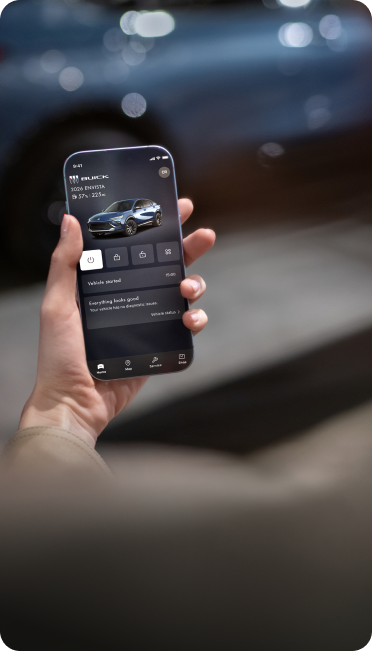 A person holds a smartphone displaying a Buick mobile application with vehicle information while standing near a parked Buick vehicle.