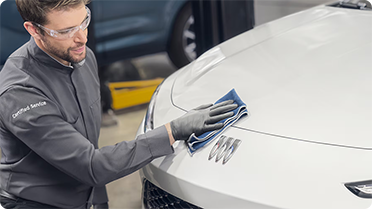 A certified service technician in safety glasses gently wipes the hood of a white Buick vehicle, revealing the distinct Buick logo.