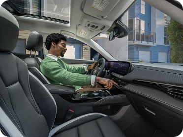 A smiling man in a green sweater sits in the driver's seat of a Buick vehicle, adjusting controls on the center console.