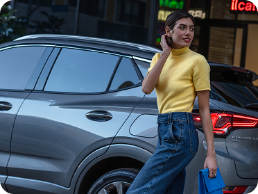 A woman in a yellow top stands near a grey Buick Encore GX parked on an urban street.
