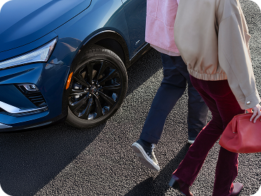 Two people walk past the front wheel and blue body of a Buick vehicle.