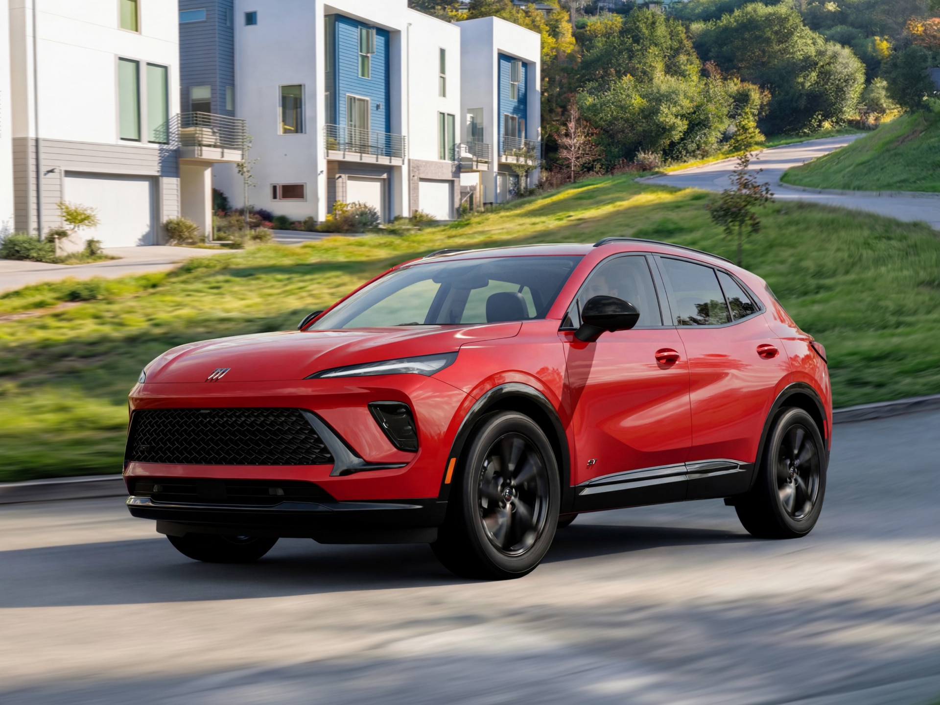 A Red Buick Envision SUV Driving on a Paved Road with Modern Houses and Green Hills in the Background