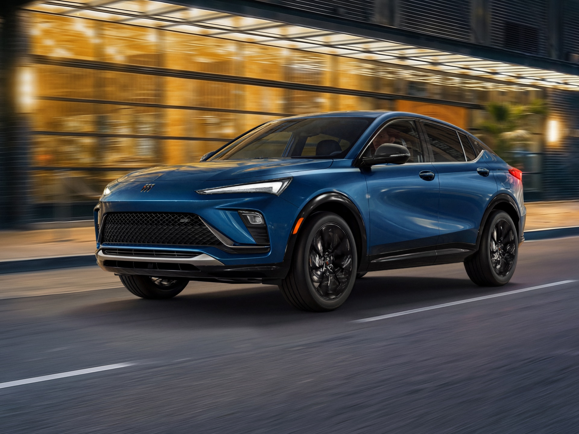 A Blue Buick Envista SUV Driving on a City Street at Dusk with Blurred Building Lights in the Background