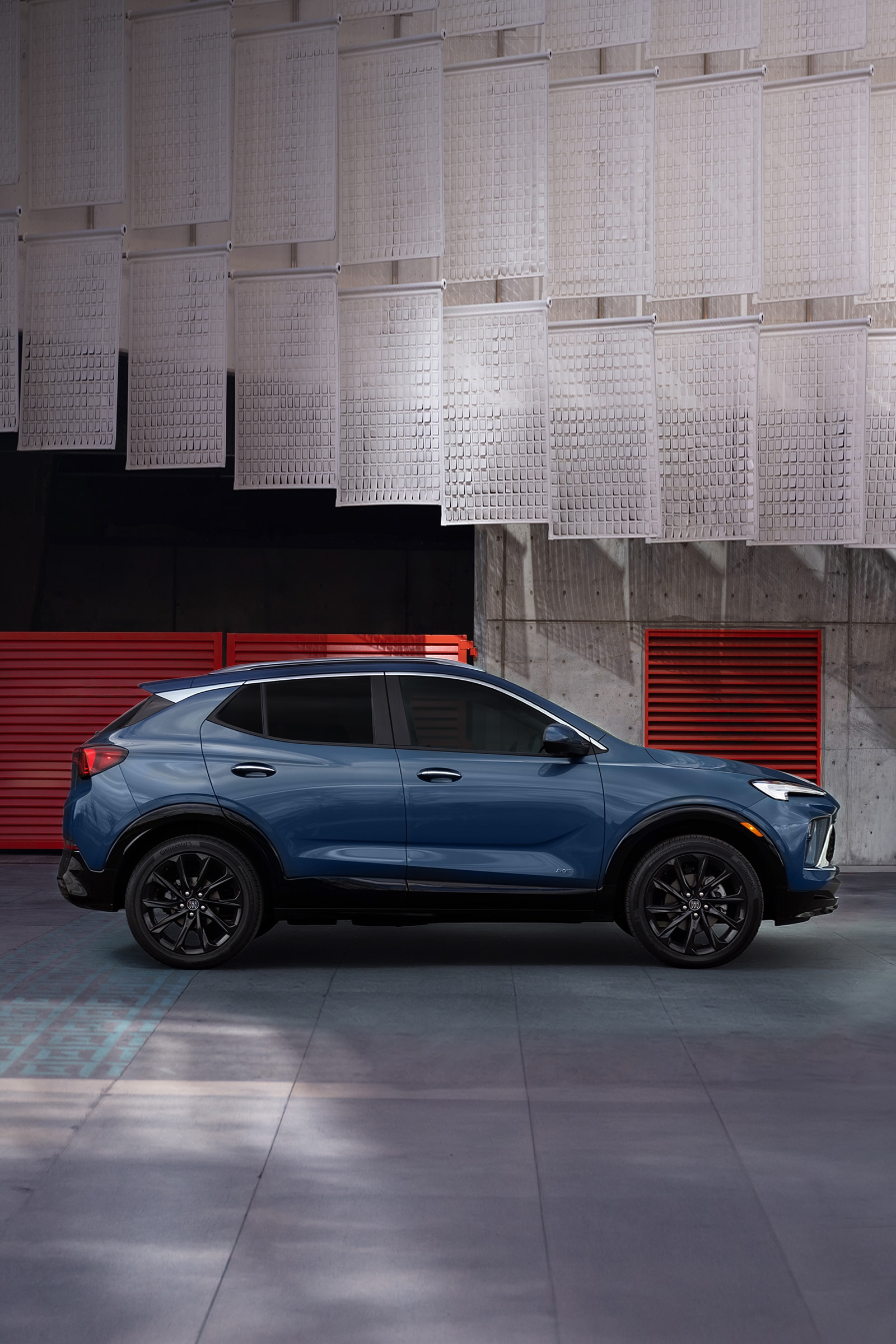 A Deep Blue Buick Encore GX SUV Is Parked Sideways with Black Wheels in Front of a Modern Building with Red Louvers